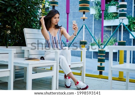 Asian hipster girl shooting video vlog via application on cellphone technology, youthful female blogger connecting to 4g on smartphone gadget for clicking selfie photos during leisure in street cafe