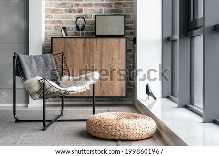 Cozy lounge zone near panoramic window with metal chair, straw ottoman and modern equipment for listening music on commode against brick wall. Loft style interior design in hotel or home Royalty-Free Stock Photo #1998601967
