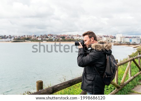 Side view of stylish adult man traveler in casual clothes and headband taking photo of rippling sea on camera standing on embankment with wooden fence under cloudy sky