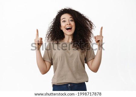 Cheerful brunette girl laughing happy, pointing fingers up and showing banner promotional text on empty space, standing against white background