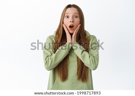 Girl drop jaw and gasp, hold hands on face fascinated, look in awe at advertisement, check out something cool, wear casual oversize hoodie, stand over white background