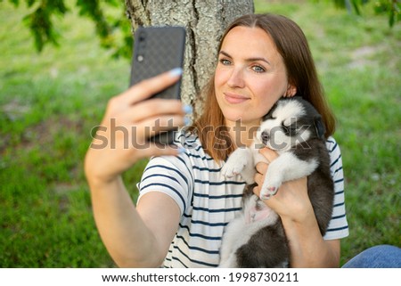 Young beautiful woman in a t-shirt and jeans with a husky puppy takes a photo or selfie on the phone while sitting in a park near a tree.