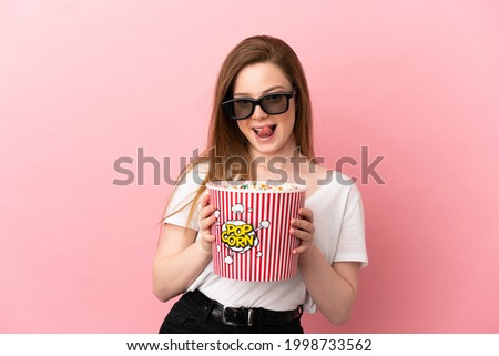Teenager girl over isolated pink background with 3d glasses and holding a big bucket of popcorns