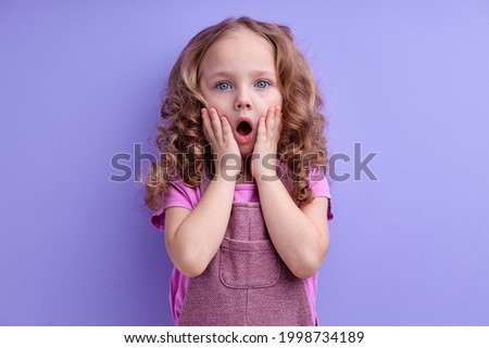 Photo of scared shocked caucasian small girl closing mouth horrified isolated on purple color background, in studio, copy space. children emotions concept