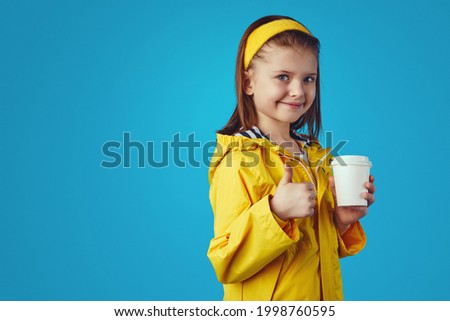 Girl wears raincoat, holds takeaway cup with tea and showing thumb up