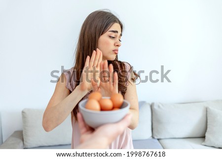 Photo of a woman refusing to eat eggs. Egg free affected allergy banned restriction. Young beautiful girl holding fresh egg at home with open hand doing stop sign with serious and confident expression