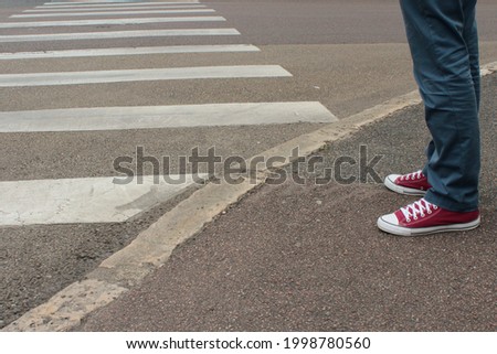 A man in blue pants and purple sneakers with white shoe laces waits on the sidewalk by the zebra pedestrian road crossing.