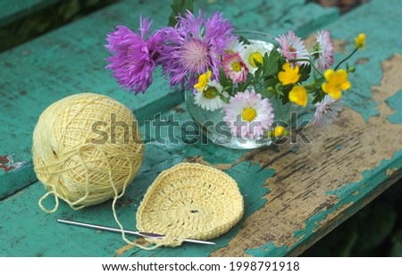 Crochet with yellow threads, lying on the table on the table, next to a standing vase with wildflowers 