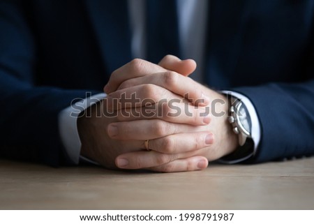 Clasped hands of businessman in formal suit sitting at work or meeting table. Attentive professional taking part in interview, negotiation, ready for dialogue, conversation. Cropped close up Royalty-Free Stock Photo #1998791987