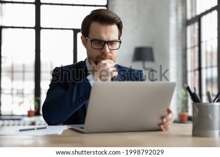 Thoughtful serious businessman reading email message on laptop screen, thinking over problem solving and making decision, watching online video conference or learning webinar, analyzing market Royalty-Free Stock Photo #1998792029