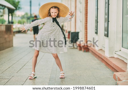 Beautiful emotional girl in elegant wide-brimmed straw hat and polka-dot dress poses on a city street. Kid's fashion. 