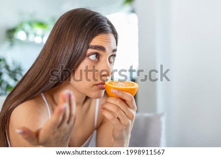 Sick woman trying to sense smell of half fresh orange, has symptoms of Covid-19, corona virus infection - loss of smell and taste. One of the main signs of the disease. Royalty-Free Stock Photo #1998911567