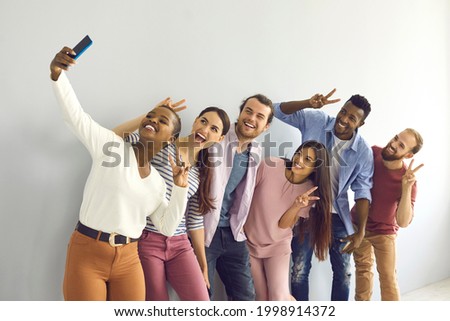 Group of smiling multinational students or friends showing v-sign gestures making selfie. African American girl is photographed with her friends using a phone while standing in a room near a wall.
