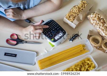 Top view female hands use labeler electronic device for marking different pasta closeup POV shot. Woman creating letters, names, titles, labels printing stickers on label machine. Household storage