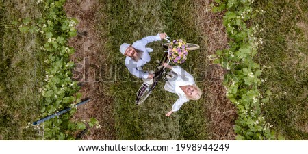 aerial drone shot, pic of blonde woman with white dress and her boyfriend or husband posing with bicycle with beautifully decorated flower basket in nature, nature concept, flowers, delivery service