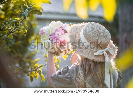 Beautiful blonde in a green summer garden. A girl in a straw hat with a bouquet of white peonies and pink carnations.