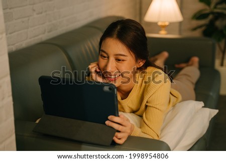 Young Asian woman relaxing at home, lying on couch on stomach with pillows using digital tablet and enjoying movies at home during late night