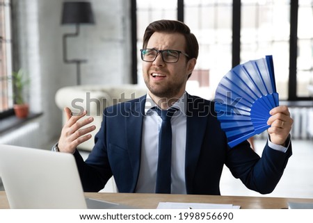 Exhausted male office employee suffering from heat, hot stuffy air. Business man cooling with handheld fun while working on summer day in office without conditioner. Conditioning, ventilation concept Royalty-Free Stock Photo #1998956690