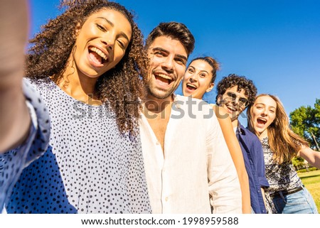 Five young multiracial friends in line posing for photo portrait smiling with open mouth. Black Hispanic brunette girl taking a selfie with smartphone outdoor. POV of happy millennial carefree people