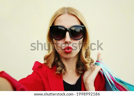 Portrait close up of young woman taking a selfie picture by smartphone with shopping bags, female model blowing her lips with red lipstick sending sweet air kiss