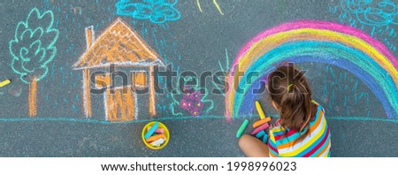 The child draws a house and a rainbow on the asphalt with chalk. Selective focus. Kids. Royalty-Free Stock Photo #1998996023