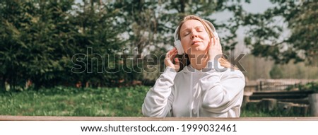 A girl in a white hoodie listens to music with headphones and relaxes sitting on a bench near a country house in nature, ASMR concept