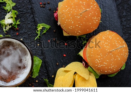 Homemade burgers on black slate dish with peppercorns, lettuce leaves, chips and glass of beer on the table, close up