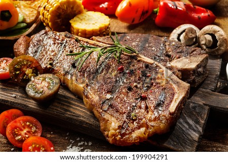 Close up of a succulent tender grilled porterhouse steak seasoned with pepper and rosemary on a wooden board with fresh halved tomatoes, mushrooms, corncobs and bell peppers