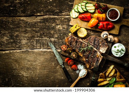 Overhead view of colorful roast vegetables, savory sauces and salt served with grilled t-bone steak on a rustic wooden counter in a country steakhouse