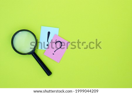 Top view photo of a magnifying glass and sticker with question mark and an exclamation mark lie on a green background.