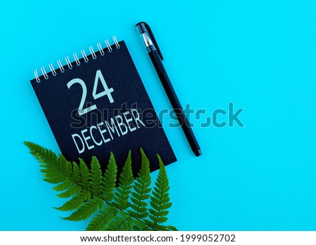 December 24th. Day 24 of month, Calendar date. Black notepad sheet, pen, fern twig, on a blue background. Winter month, day of the year concept.