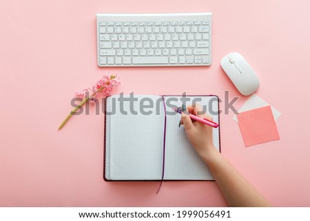 Pink work desk with female hand writes notes in notebook at workplace. Desktop office space top view. Work space girly office table. Back to school layout concept.