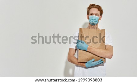 Picture of a young redhead courier hugging paper bag and and wearing blue medical mask with rubber gloves . The most safe delivery in the world High quality photo