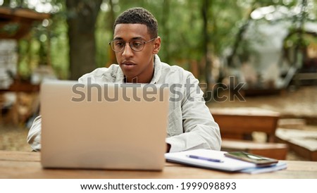 Young african american freelancer working on laptop while sitting at wooden table outdoors, widescreen. Work in nature concept