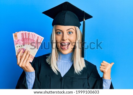 Beautiful blonde woman wearing graduation cap and ceremony robe holding indonesian rupiah pointing thumb up to the side smiling happy with open mouth 