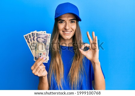 Young hispanic woman wearing delivery uniform and cap holding japanese yuan doing ok sign with fingers, smiling friendly gesturing excellent symbol 