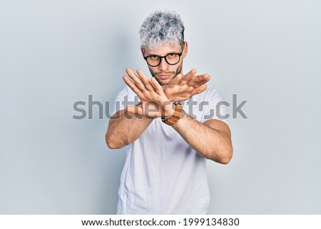 Young hispanic man with modern dyed hair wearing white t shirt and glasses rejection expression crossing arms and palms doing negative sign, angry face 