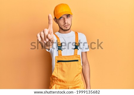 Hispanic young man wearing handyman uniform pointing with finger up and angry expression, showing no gesture 