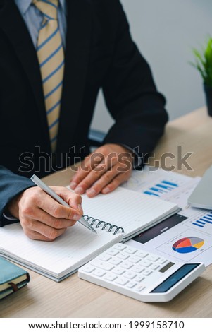 Businessman holding pen and pointing paper chart summary analyzing annual business report with using laptop at the room office desk. Financial reporting and results
