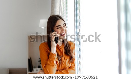Smiling business woman standing and leaning on a glass wall, talking on a smart phone, copy space. Portrait of smiling beautiful business woman, looking aside and talking on mobile in light office.
