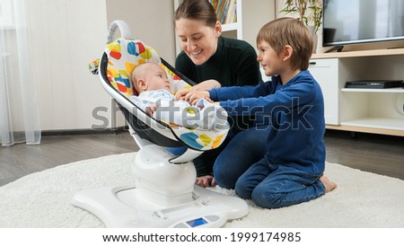 Older brother and smiling mother playing with little baby boy rocking in electric rocking seat. Child development and happy childhood Royalty-Free Stock Photo #1999174985