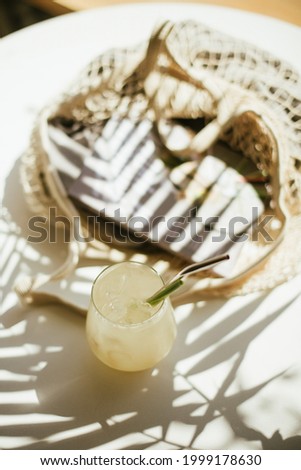 Delicious lemonade in a glass on the white cafe table with string bag and a magazine inside, with palm shadows on it, top view
