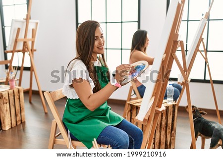 Young hispanic artist women painting on canvas at art studio smiling with happy face winking at the camera doing victory sign with fingers. number two. 