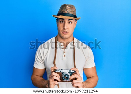 Hispanic young man holding vintage camera in shock face, looking skeptical and sarcastic, surprised with open mouth 