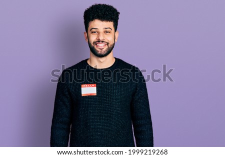 Young arab man with beard wearing hello my name is sticker identification looking positive and happy standing and smiling with a confident smile showing teeth 