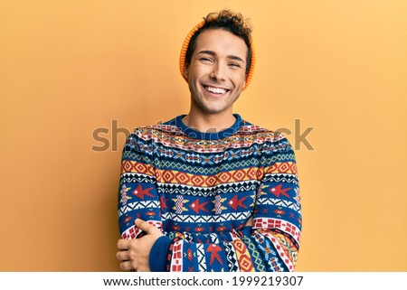 Young handsome man wearing wool hat and colorful sweater happy face smiling with crossed arms looking at the camera. positive person. 