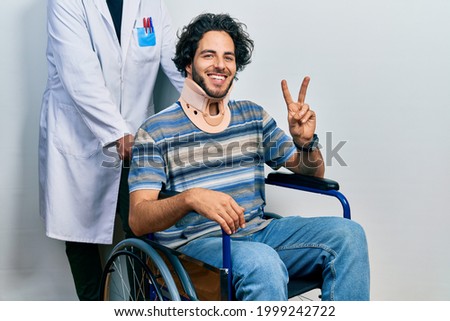 Handsome hispanic man sitting on wheelchair wearing neck collar showing and pointing up with fingers number two while smiling confident and happy. 