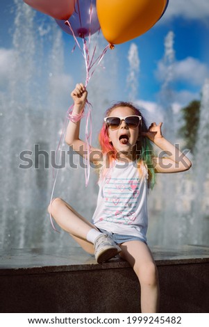 cute caucasian smiling girl with colorful dyed hair wearing mom's sunglasses holding baloons sitting near fountain. Image with selective focus