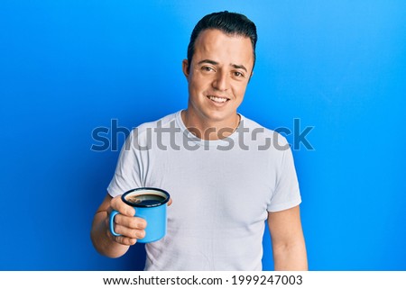 Handsome young man drinking a cup coffee looking positive and happy standing and smiling with a confident smile showing teeth 