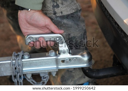 A man's hand checks the fixation of the trailer closed hitch lock handle on the towing ball towbar of the car closeup, the safety of driving with a trailer on the road Royalty-Free Stock Photo #1999250642